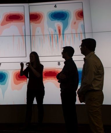 Researchers in foreground discussing plot data displayed on a large screen. 